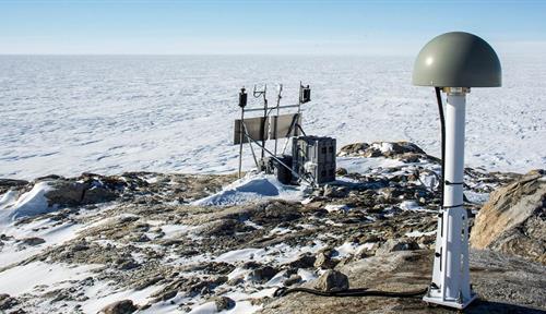 Photo shows a GPS station atop a rocky outcrop in the foreground. A snowy plain extends beyond it, under blue sky.