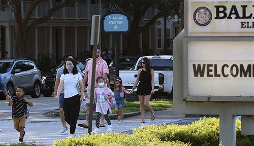 Photograph of parents and children walking toward a school. Some are wearing masks, some art not. Kids are carrying backpacks. A school crossing attendant wearing an orange vest and holding a stop sign is in the background. A large “welcome back” sign is in the foreground. 