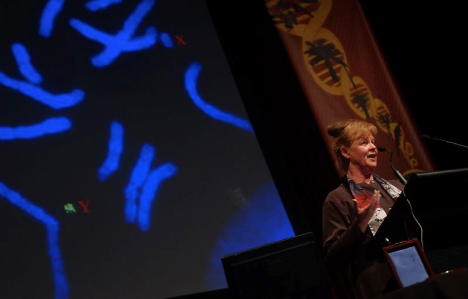 Photo of Graves standing in front of a lectern speaking into a mic. A slide of glowing blue chromosomes is projected behind her.