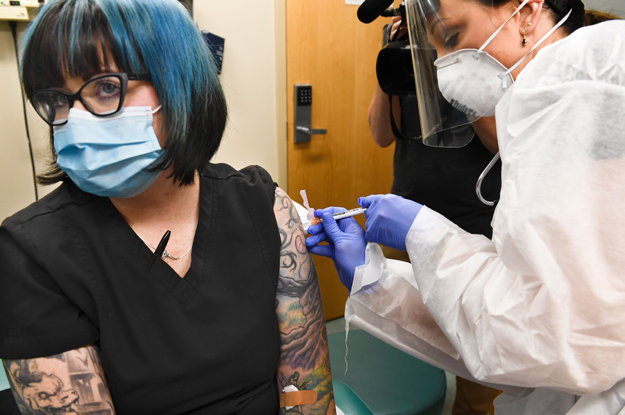Moderna vaccine trial Photograph of a woman wearing a black, short-sleeve top and blue surgical mask receiving an injection in her left arm by a nurse wearing blue gloves, N95 mask, face shield and white gown.