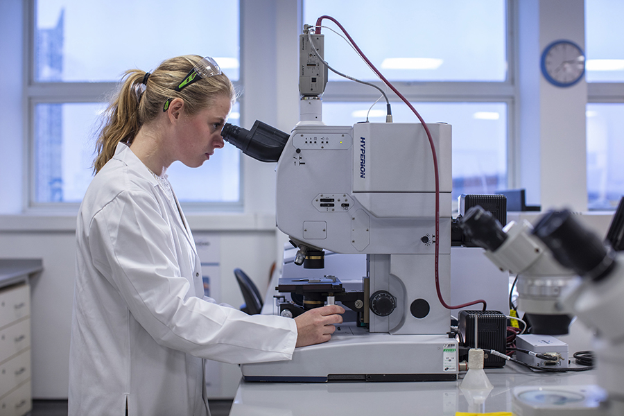 Photograph of Imogen Napper wearing white lab coat. She is looking through the eyepiece of a microscope. There are windows behind her.