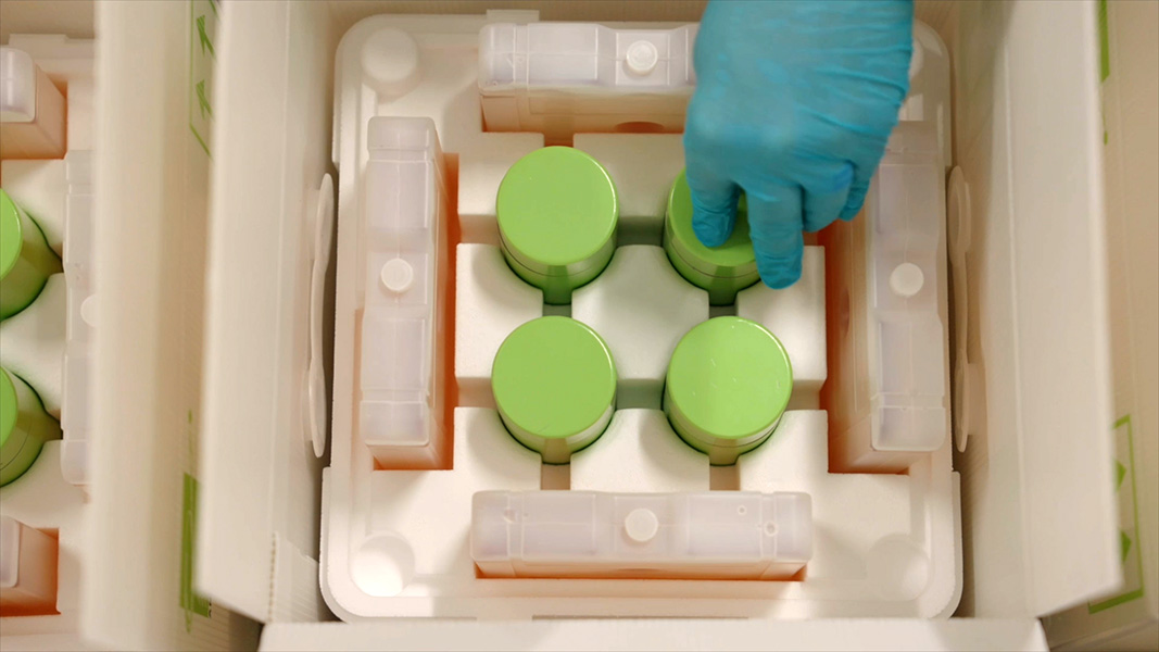 Photo shows a hand wearing a blue glove handling a round, green container — one of four within a lined box. Inside the containers is the radioactive drug.