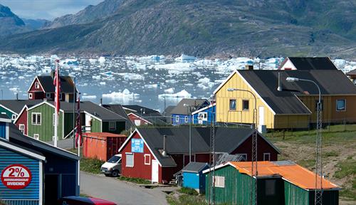 Photograph of colorful wooden houses with a bay in the background and mountains on the far shore. Chunks of ice are floating in the water.