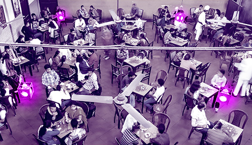 A tinted aerial photo shows a bunch of people sitting at tables in a cafeteria with lamps emitting violet light.